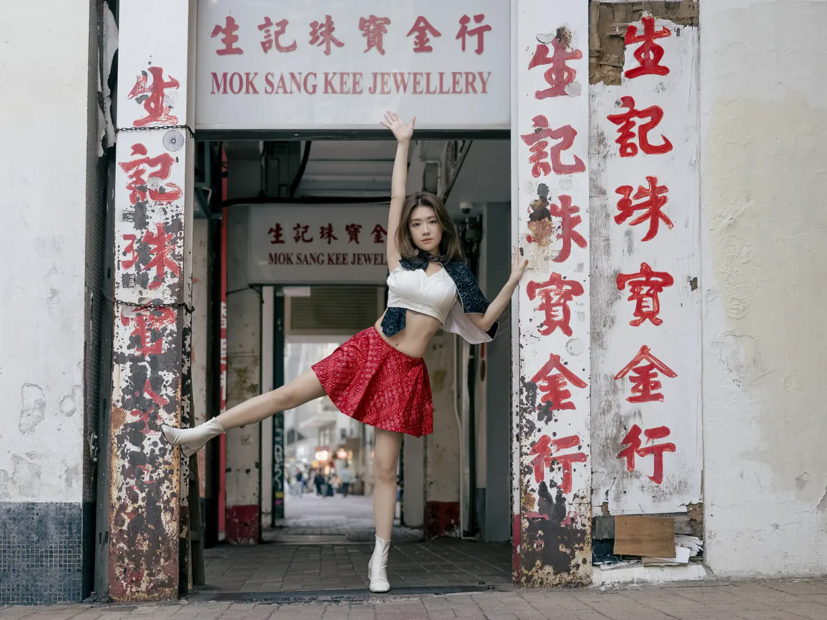 Ivy Liu balances with one arm raised and one leg extended between weathered red Chinese shop signs in Hong Kong