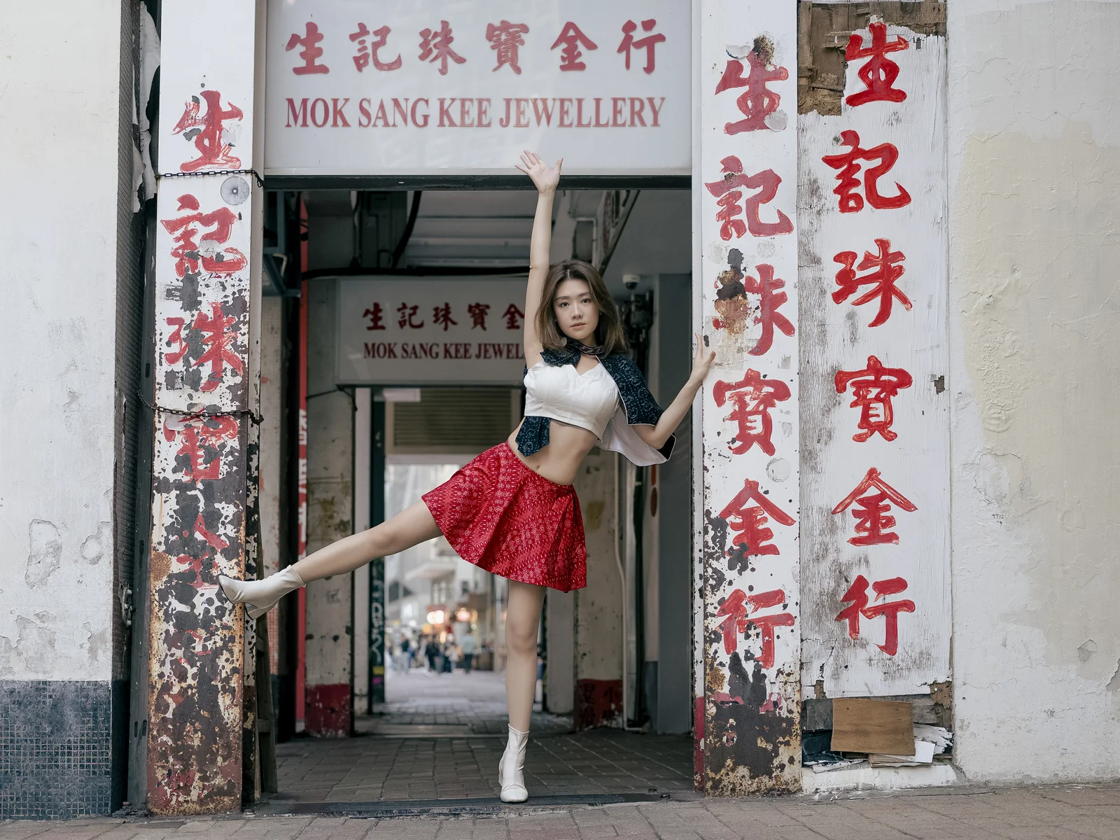 Ivy Liu balances with one arm raised and one leg extended between weathered red Chinese shop signs in Hong Kong