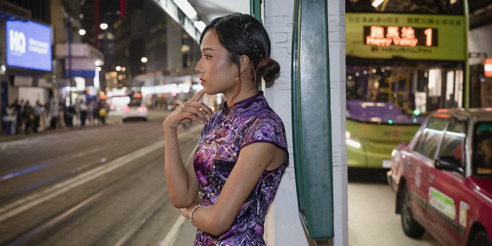 Lia stands in profile beside a tram line with a bus and city lights behind her at night