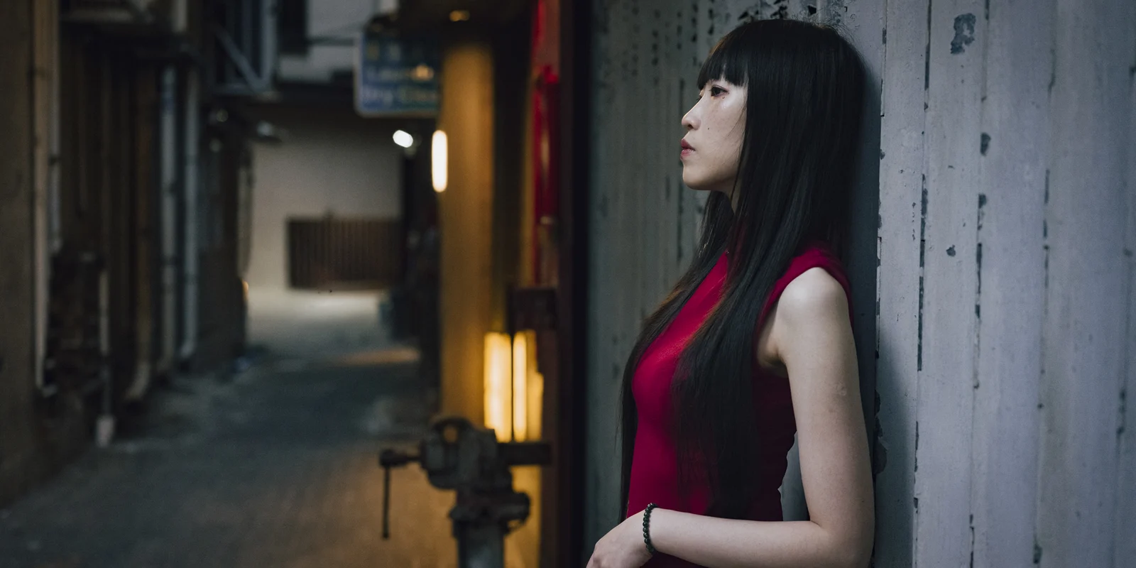 Merry Cheung in a red dress stands in profile within a dark concrete Hong Kong alley