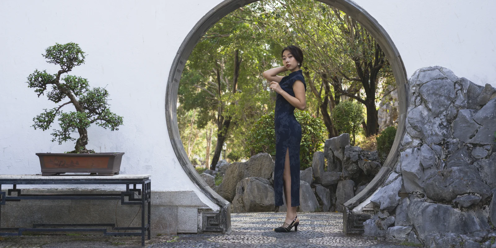 Chow stands inside a circular garden opening framed by rock, white wall, and green trees