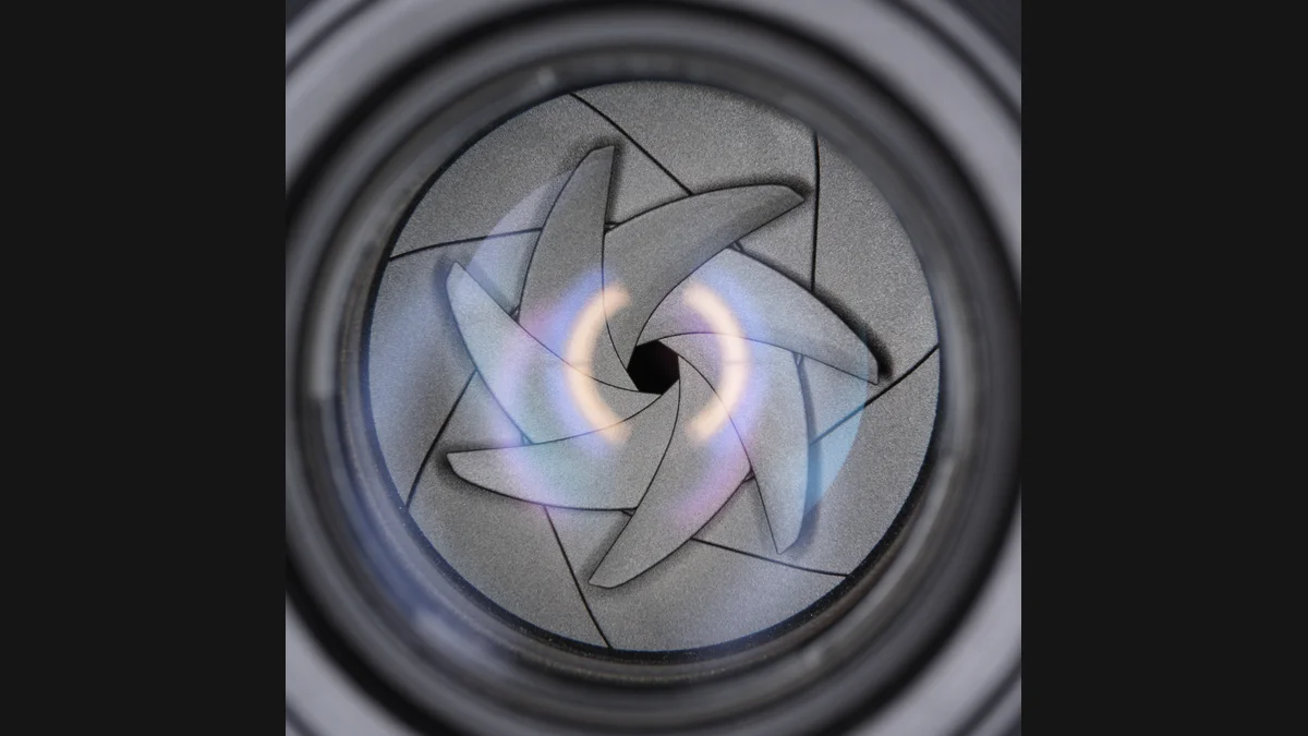 A photograph of overlapping aperture blades forming a small opening inside a lens, preserved with dark side matting