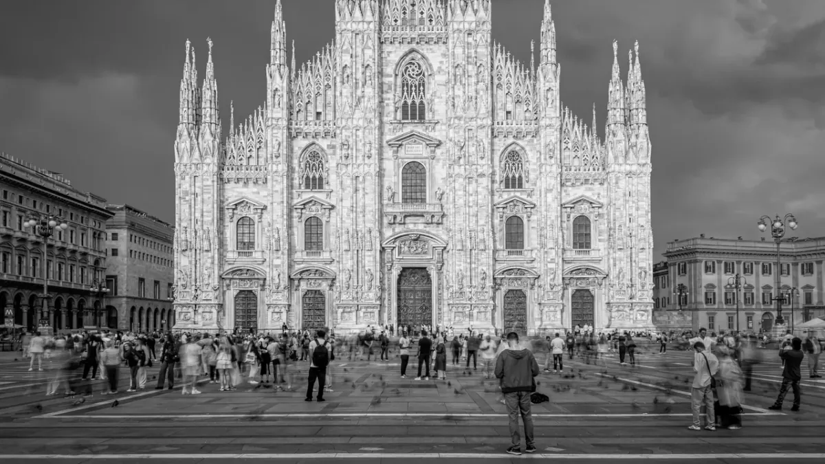 Black-and-white photograph of Milan Cathedral with blurred moving visitors and distinct stationary figures in the square.