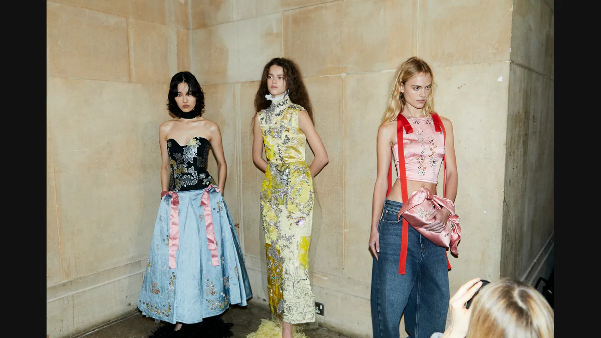 Three models in embellished garments against a stone wall in London Fashion Week’s official photograph