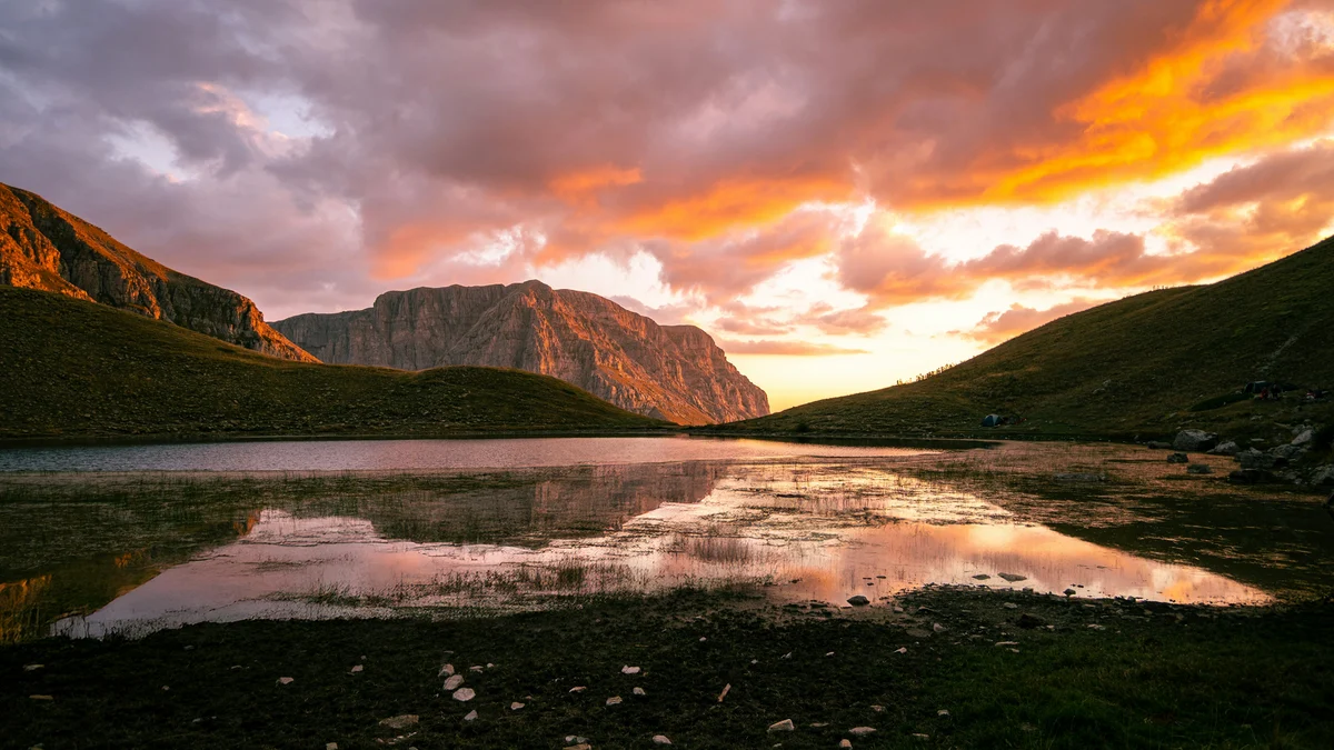 Sunset clouds glow above Dragon Lake and dark mountain slopes in Tymfi, Greece, with reflected light across the water.