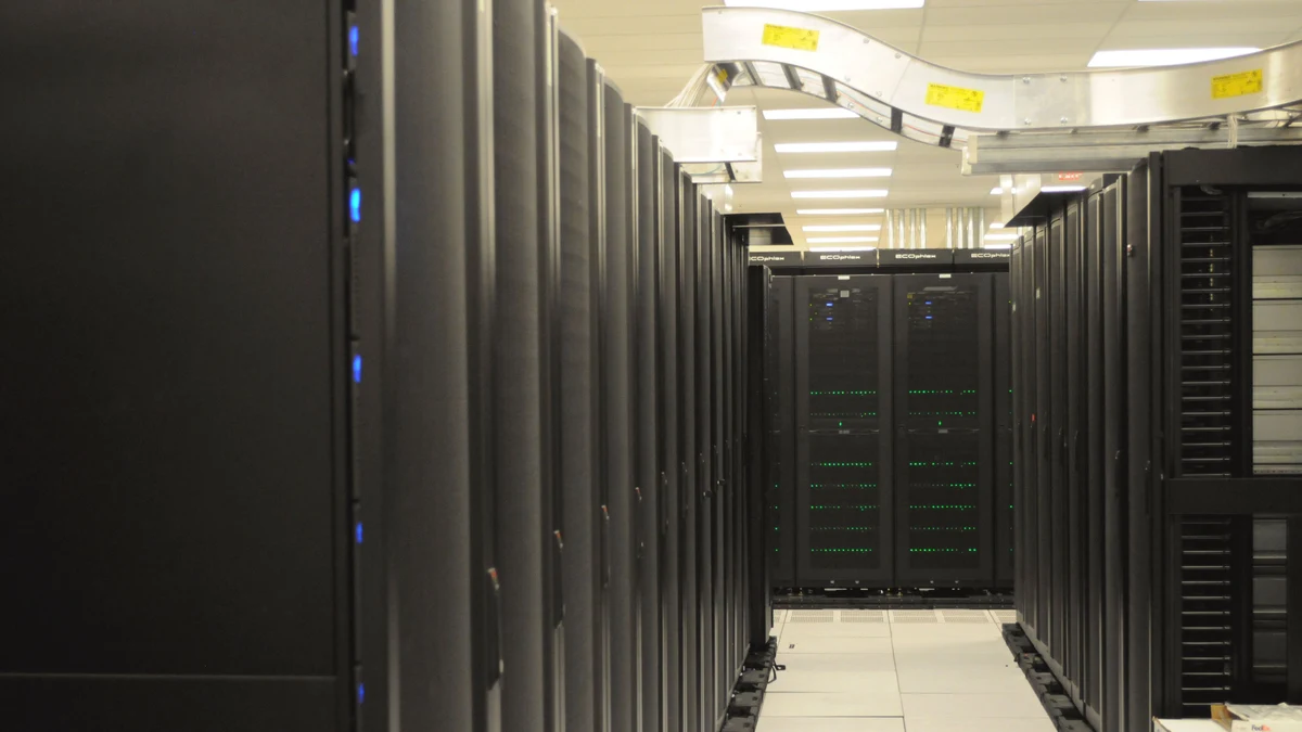 Rows of black server racks in a computing center with overhead cable trays and a bright central aisle.