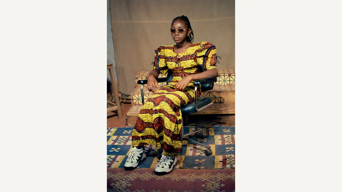 A seated sitter in dark glasses, a yellow patterned outfit, and white trainers poses in Sanlé Sory’s studio with a patterned rug and brown fabric behind the chair