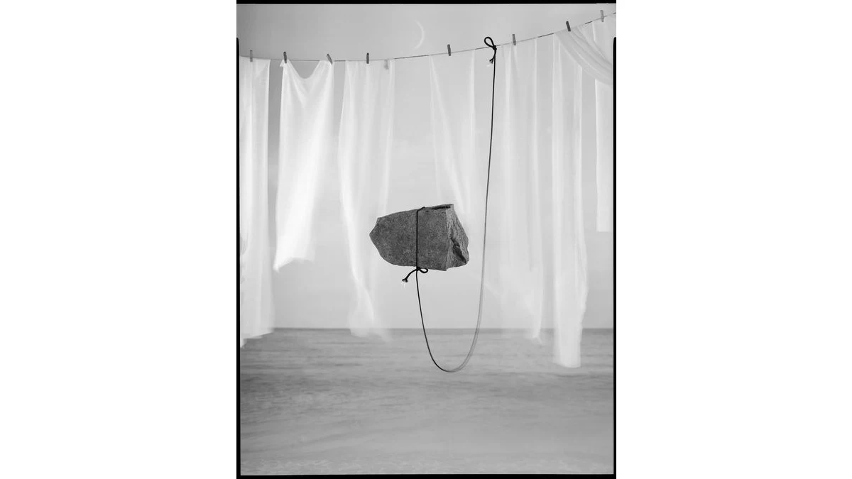 Joshua YeheNara’s black-and-white photograph of a rock apparently suspended before hanging white sheets, with a dark cord looping beneath it.